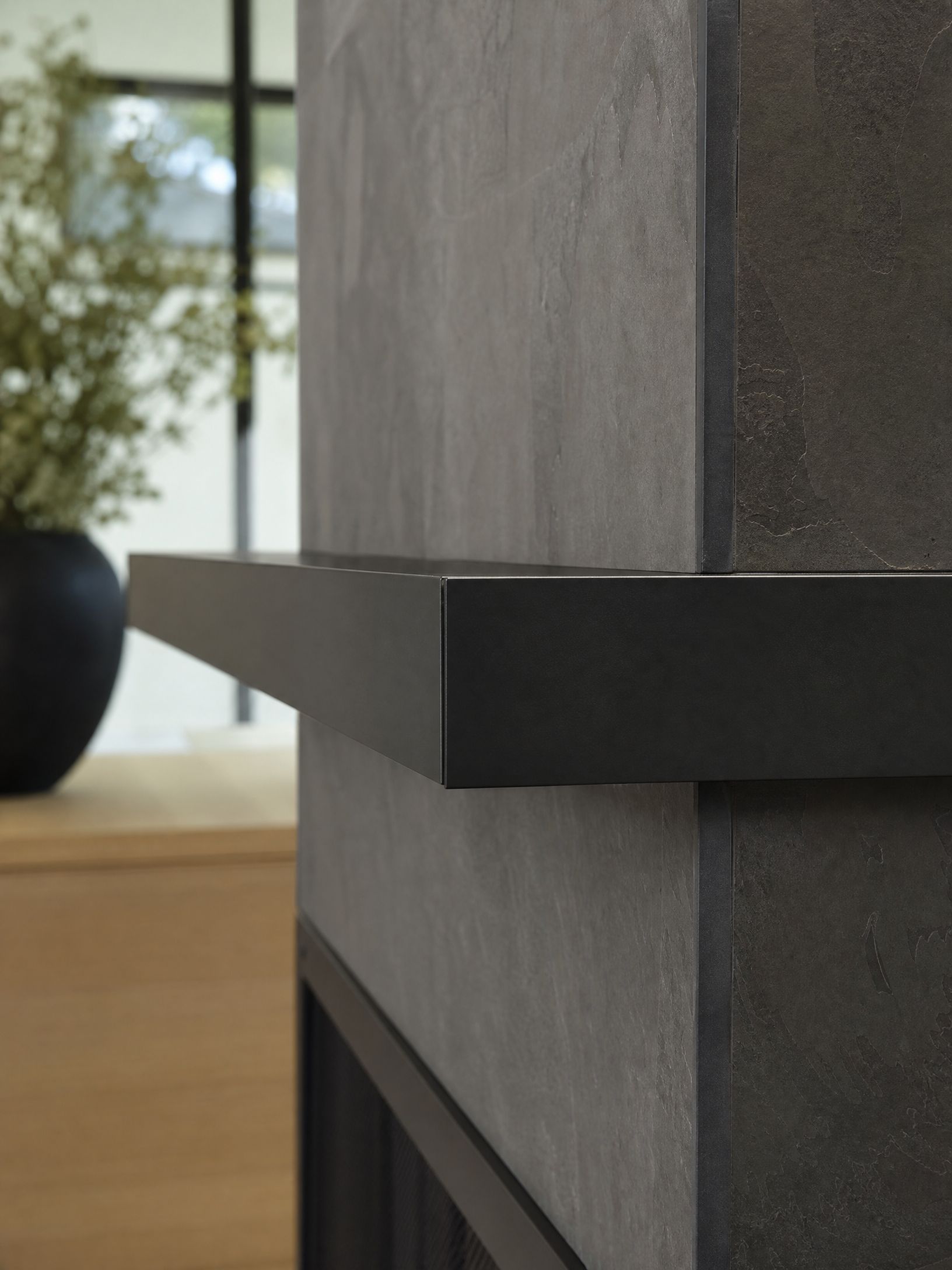Close-up of a gray stone fireplace with a black shelf against a bright windowed room