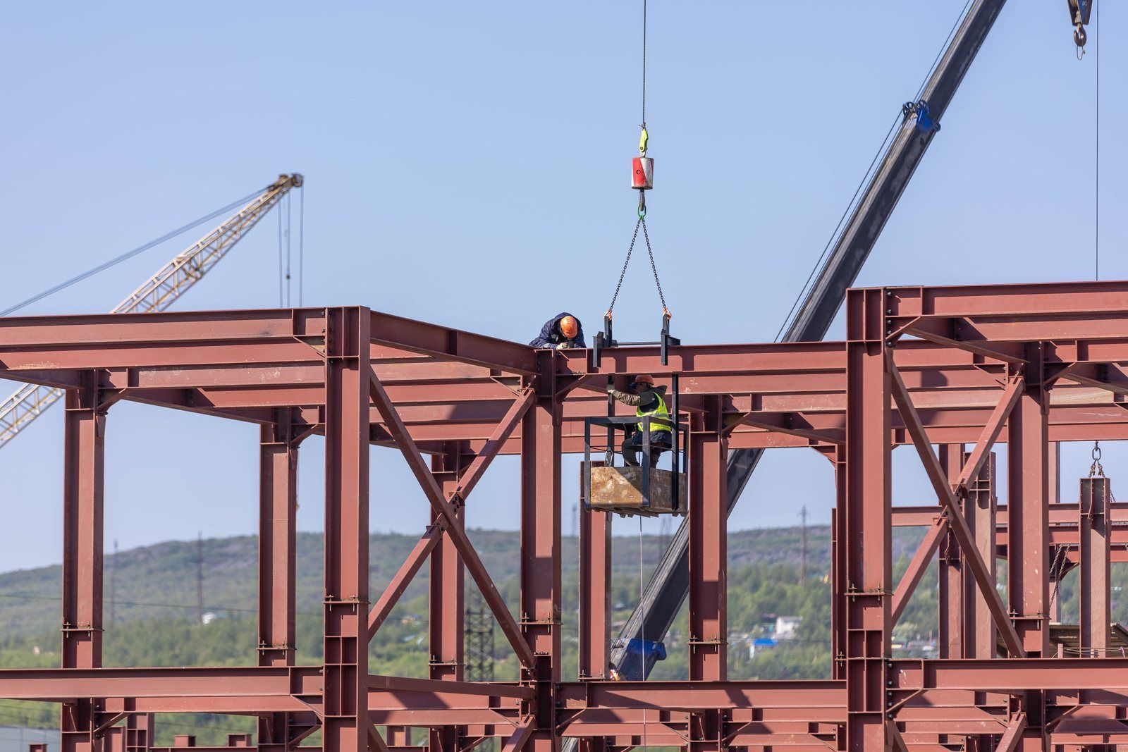 Ironworkers setting red-primed structural steel beams and columns with a crane against green hills