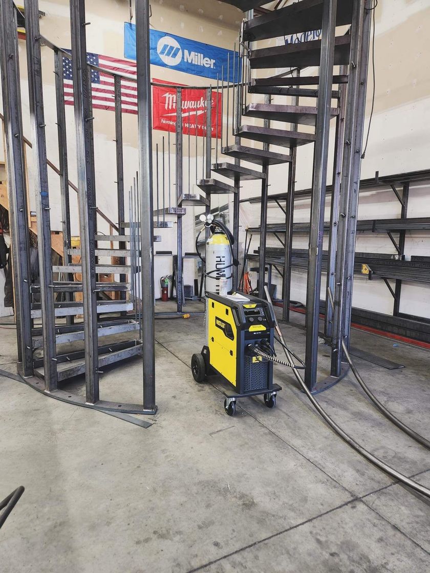 Industrial workshop with a black spiral staircase frame and a yellow welding machine on a concrete floor.