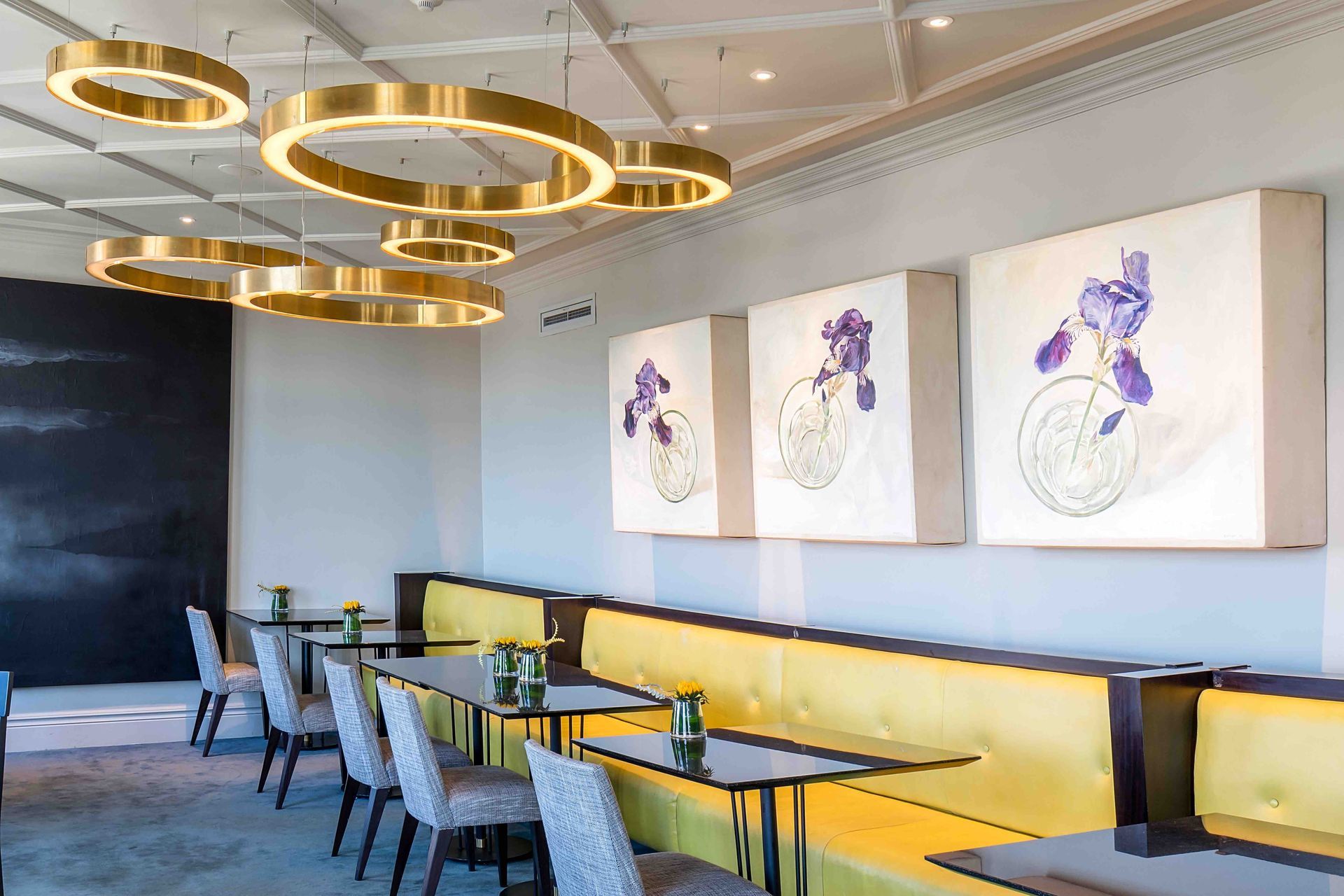 Modern cafe interior with gold ring lights, yellow banquette seating, and floral wall art