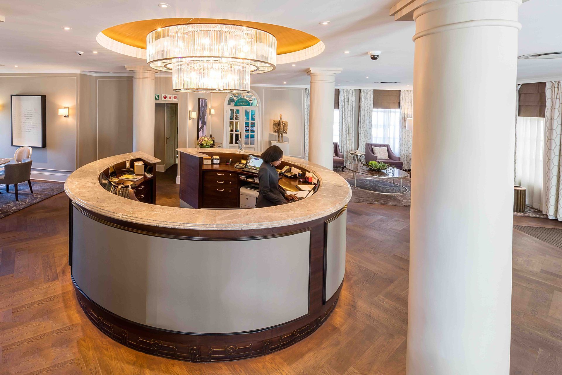 Elegant hotel lobby with a curved marble reception desk and chandelier under a bright circular ceiling.