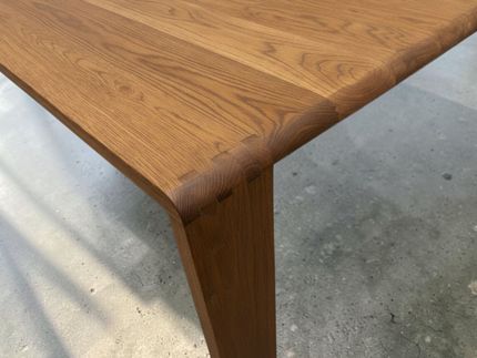 Close-up of a wooden table corner with a rounded edge and angled leg on a concrete floor