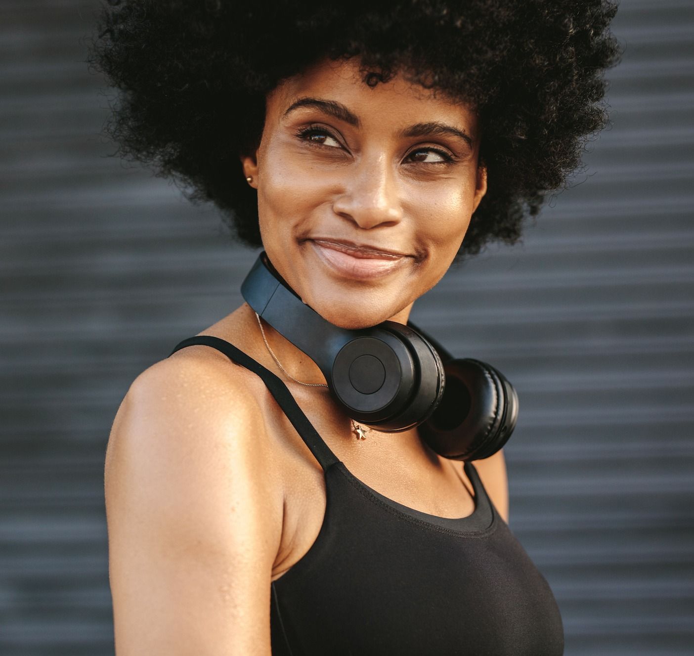 Woman with headphones around her neck, smiling, looking away from the camera. Black tank top, dark background.