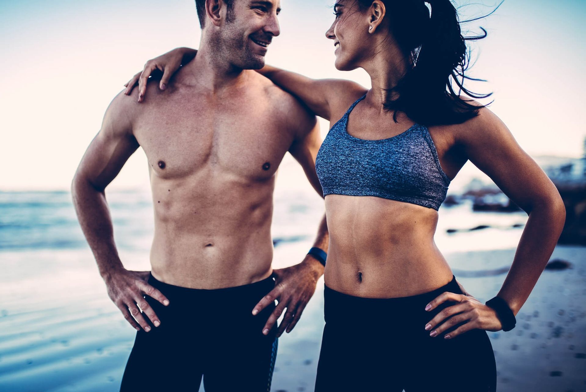 Man and woman in workout clothes on a beach, smiling and embracing after exercise.