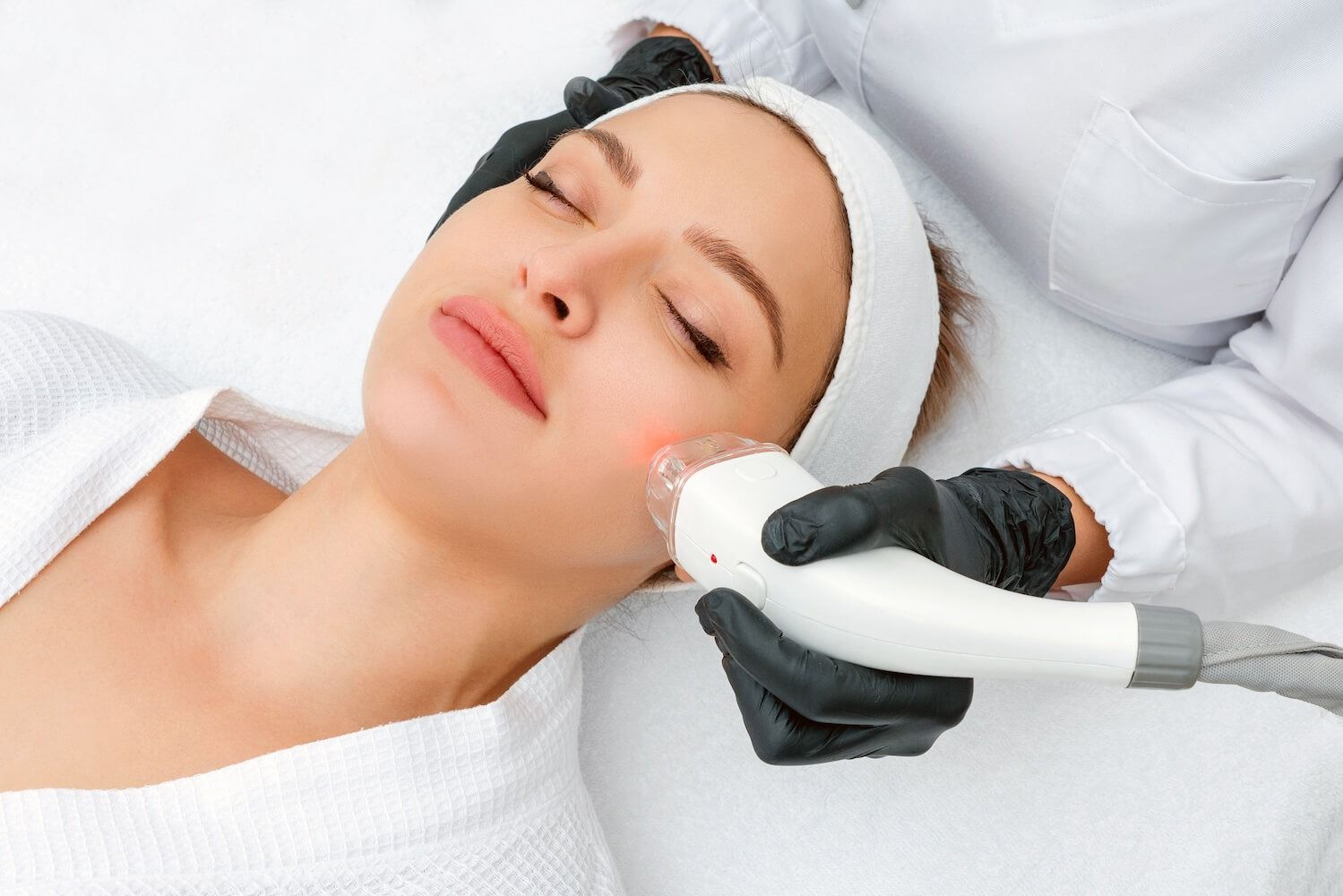 Woman receiving facial laser treatment; red light on cheek, white setting.