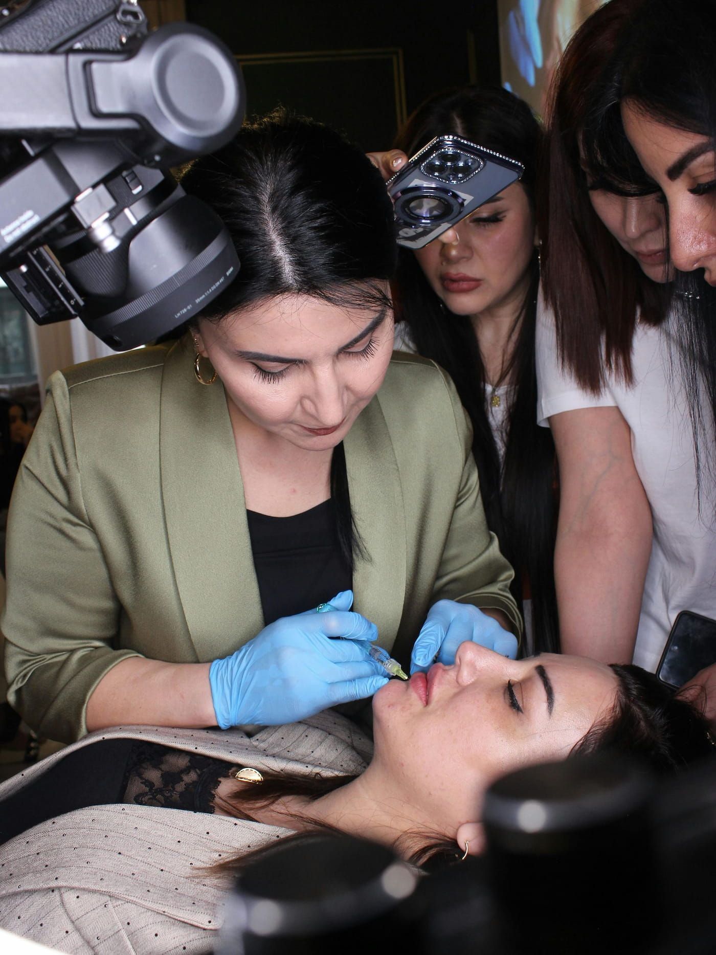 Woman in a green jacket injects lips of another person lying down. Others observe closely with lights.