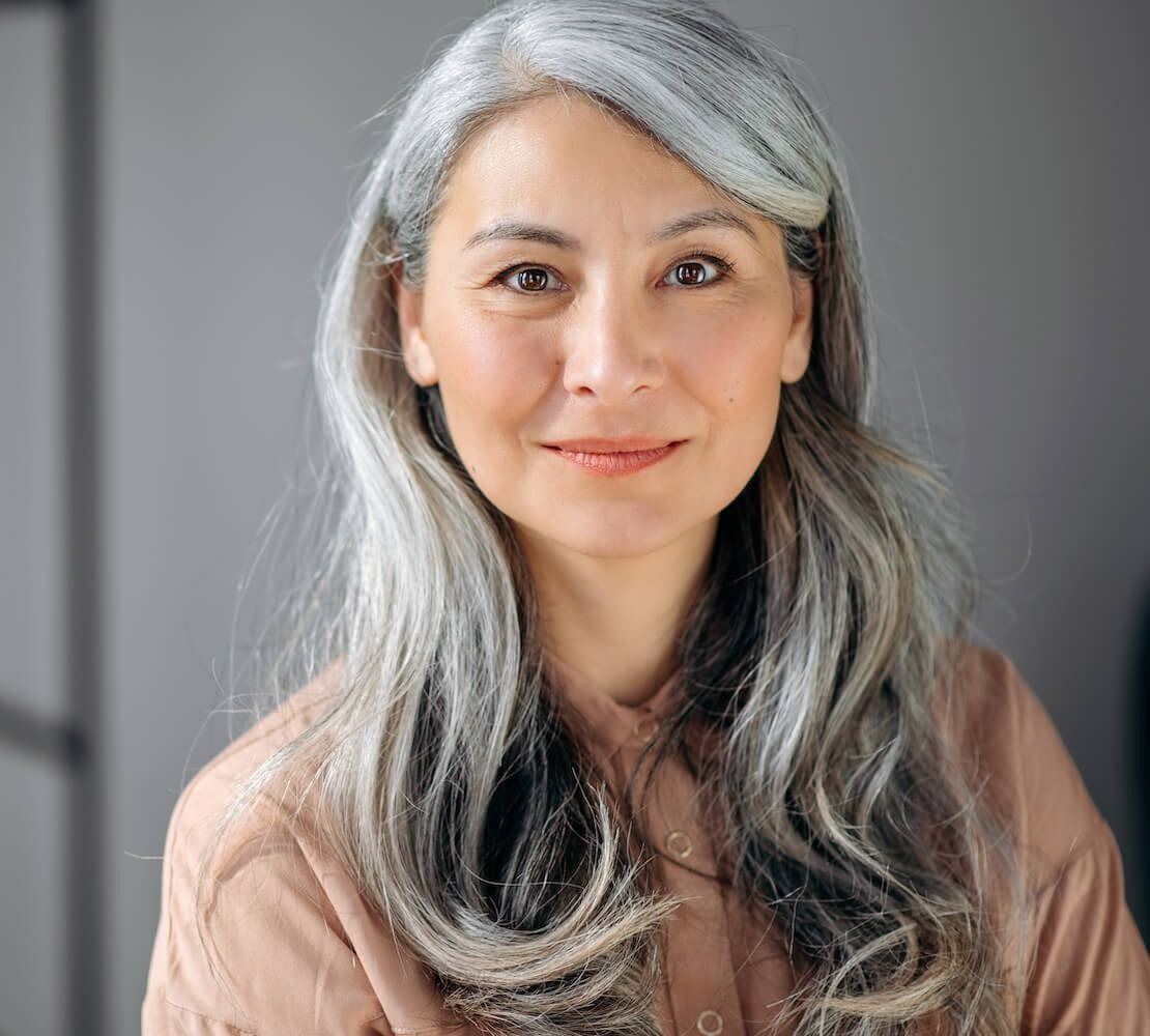 Woman with long, silver hair smiles at the camera, wearing a light brown top.