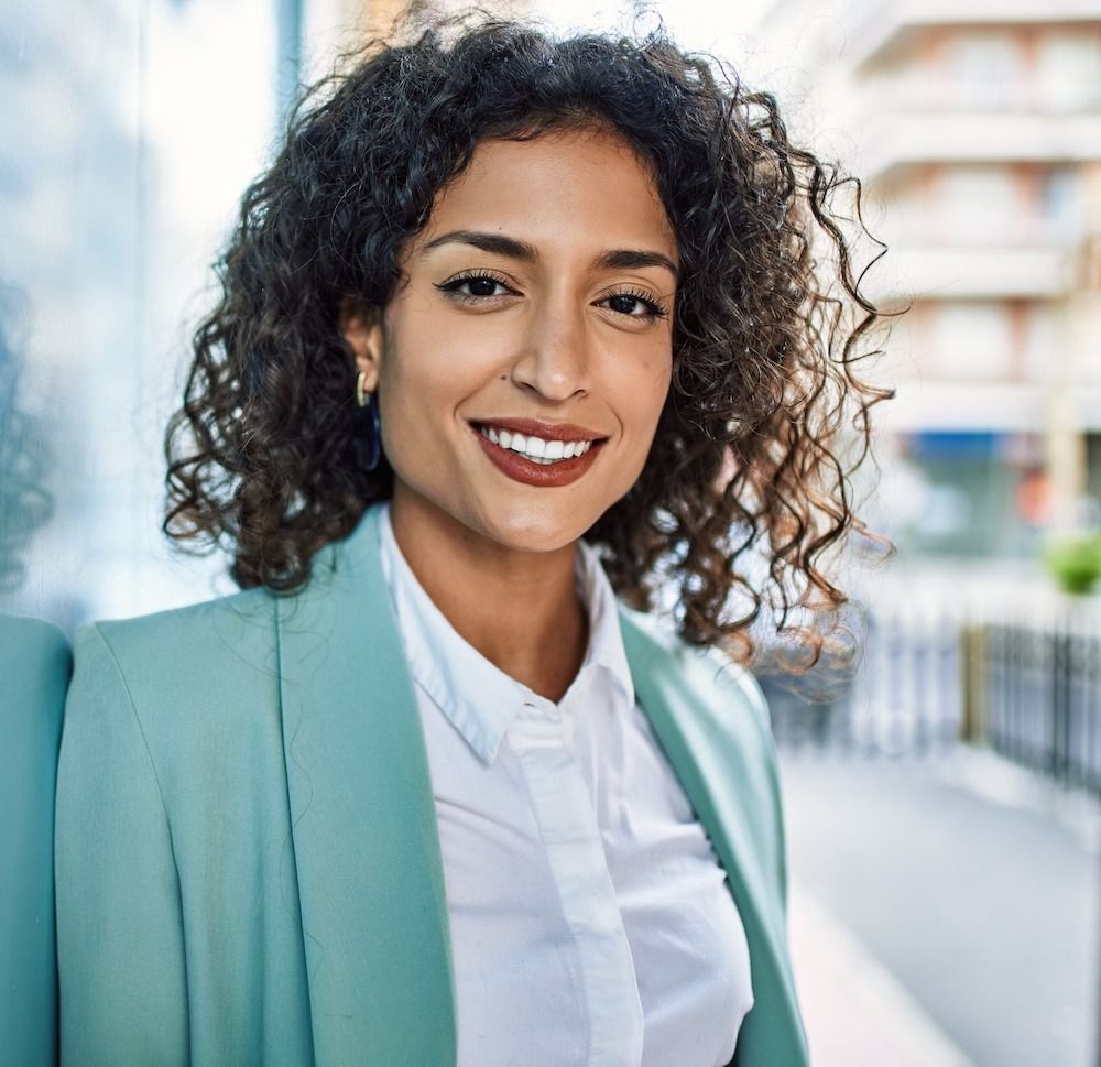 Woman in light blue blazer, smiling outdoors, leaning on a wall.