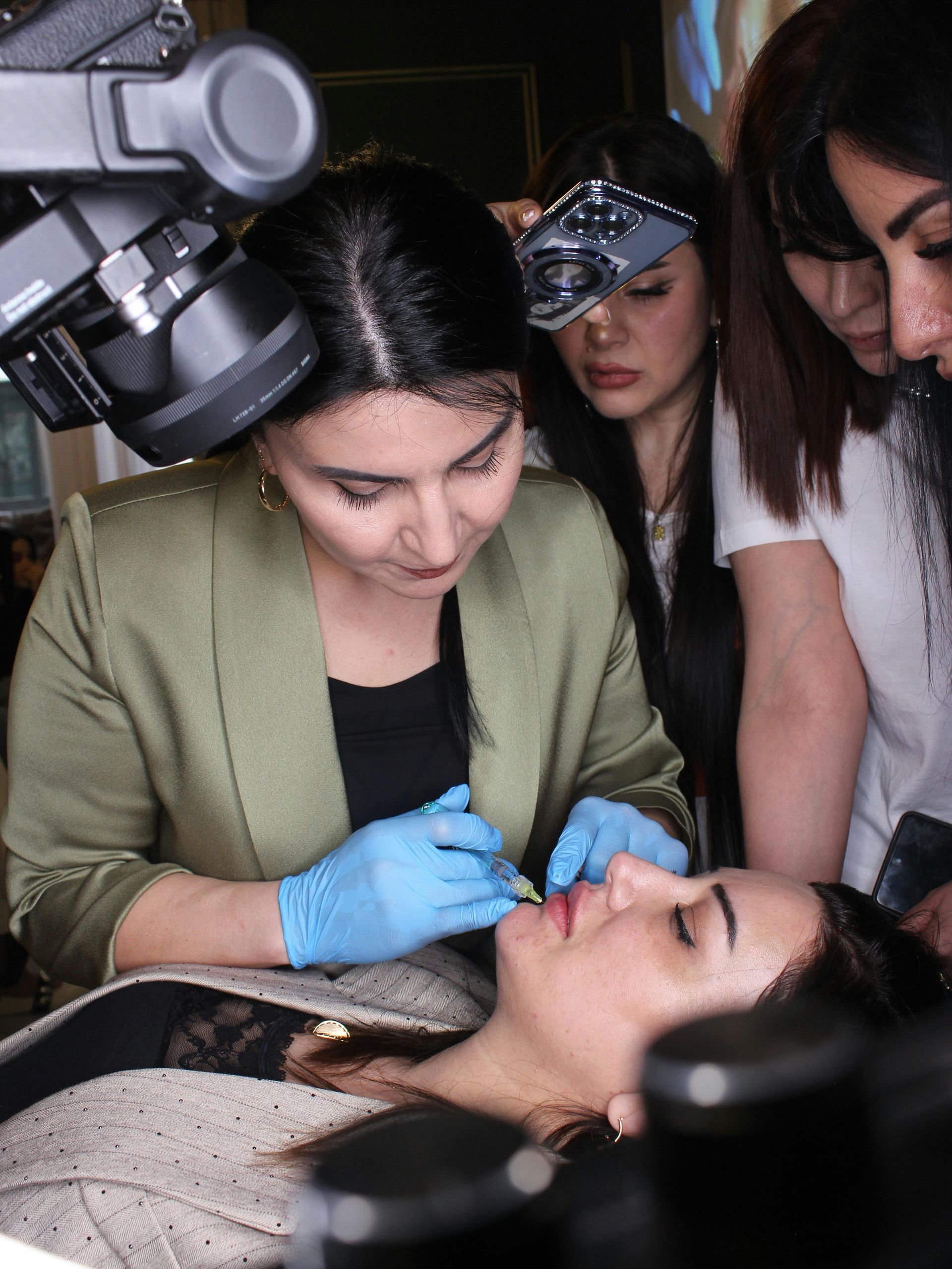 Woman in blue gloves injecting patient's lip, surrounded by observers, lit by overhead camera.