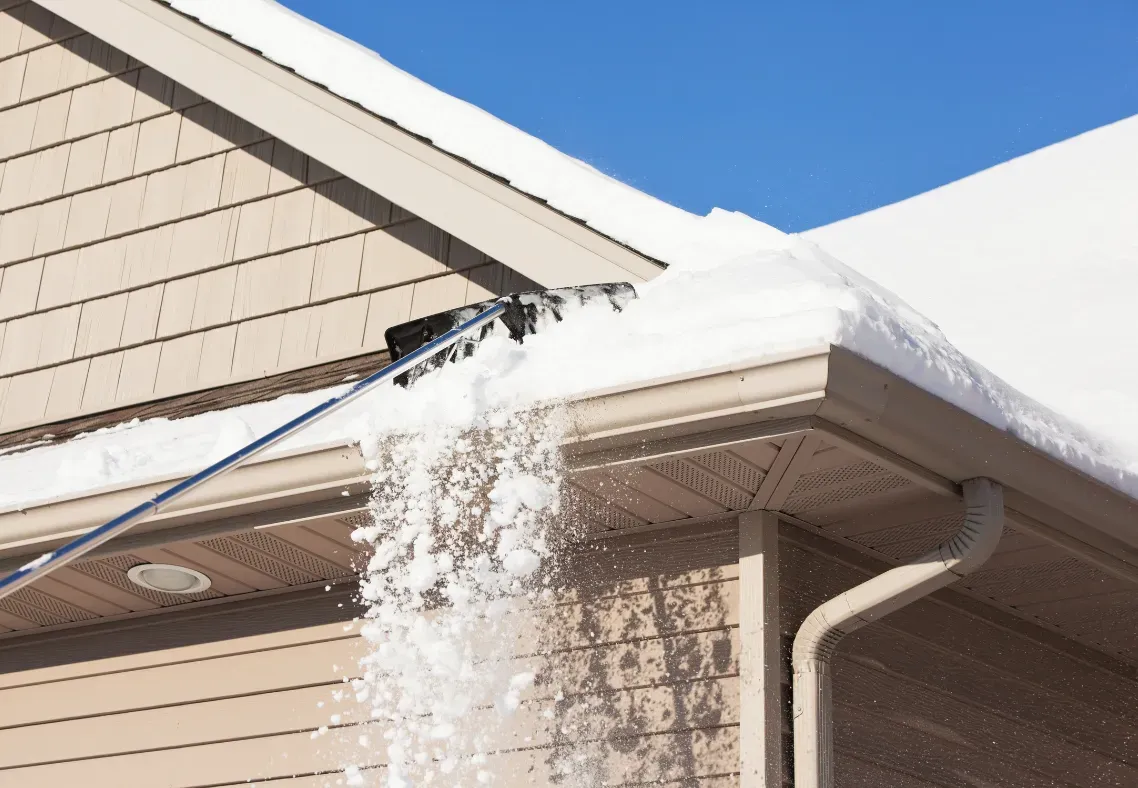 Snow being removed from a roof with a long-handled snow removal tool.