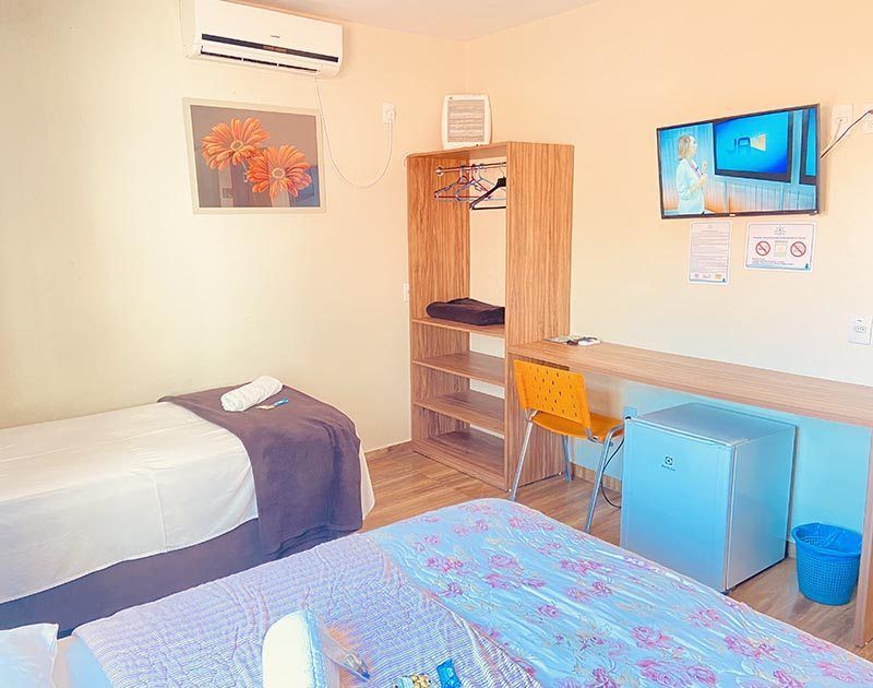 Um quarto de hotel simples com duas camas, um guarda-roupa aberto de madeira, um frigobar azul, uma escrivaninha e uma televisão na parede.