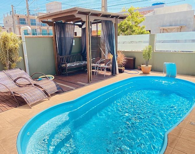 Uma piscina azul vibrante no quintal, ao lado de uma pérgola de madeira com assentos sombreados, cercada por um deck pavimentado.
