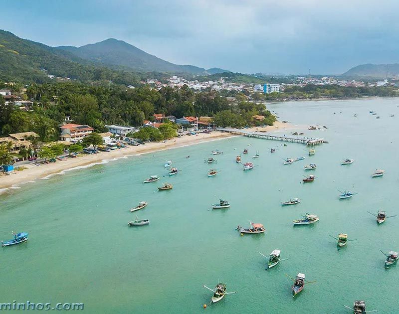 Vista aérea de pequenos barcos de pesca ancorados em águas turquesas ao longo de uma costa arenosa, tendo como pano de fundo colinas verdejantes.