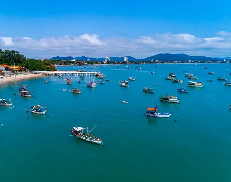 Vista aérea de vários barcos de pesca coloridos ancorados em uma baía tranquila de águas turquesa, com praia de areia e montanhas ao longe.