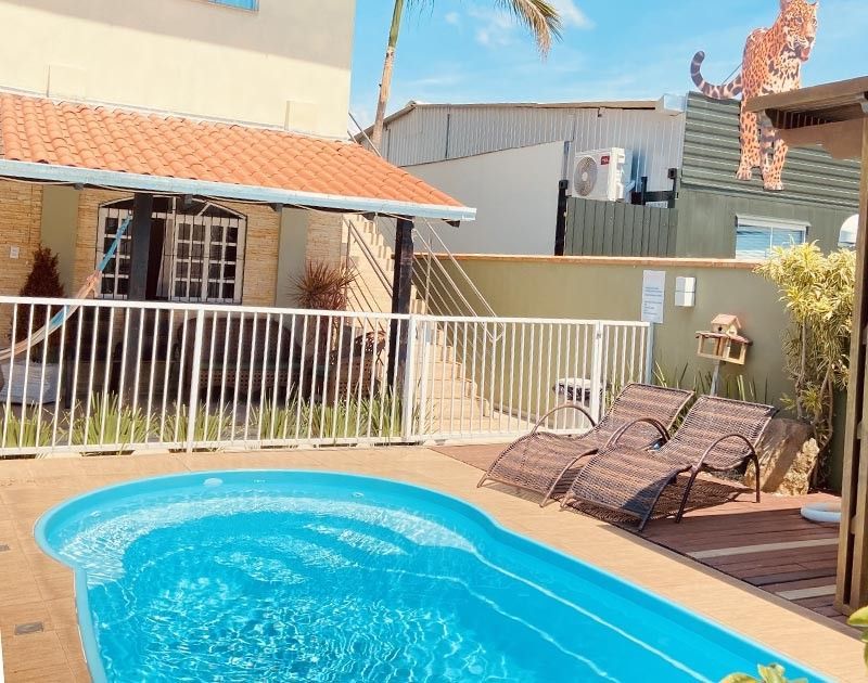 Uma piscina turquesa fica em frente a uma casa com telhado de telhas, grades brancas e duas espreguiçadeiras em um deck de madeira.