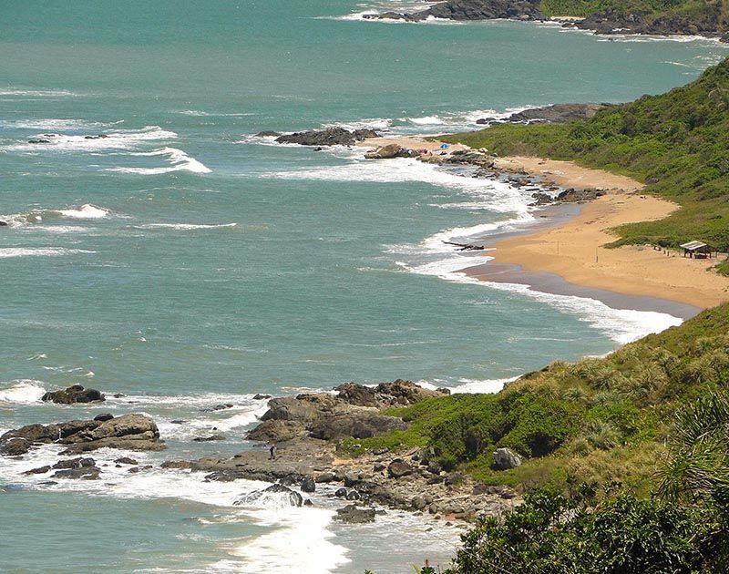 Uma vista panorâmica de uma praia de areia que encontra as águas azul-turquesa do oceano, ladeada por colinas costeiras verdejantes e rochas dispersas.