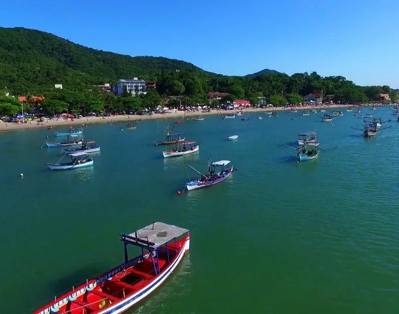Vários pequenos barcos de pesca flutuam nas águas turquesas de uma baía costeira ensolarada, com uma praia de areia e colinas ao fundo.