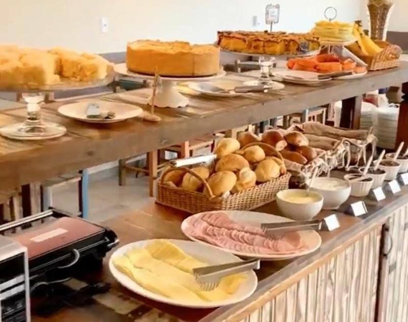 Um buffet de café da manhã com bolos, pãezinhos, presunto fatiado e queijo, servido em uma mesa rústica de madeira.
