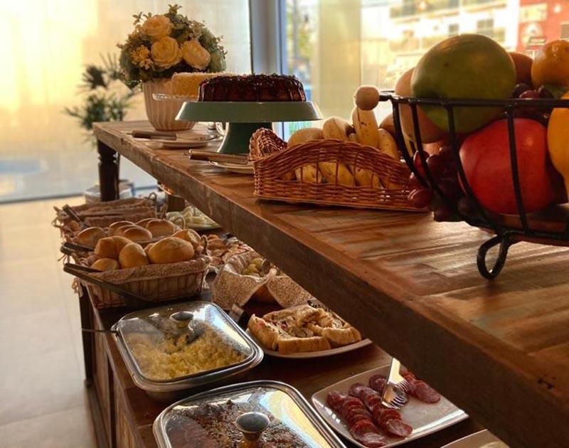 Um buffet de café da manhã com cestas de pão, frutas, bolo e bandejas de ovos mexidos e carnes sobre uma mesa de madeira.