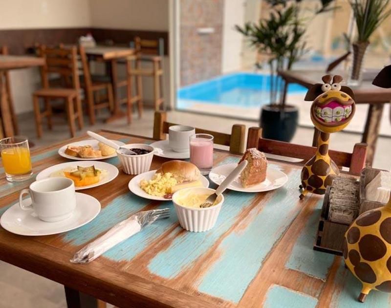Café da manhã servido em uma mesa rústica de madeira com xícaras, pratos e decorações com tema de girafa, perto da piscina.