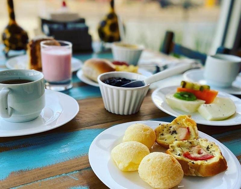 Uma mesa de café da manhã com pão de queijo, fatias de frutas, geleia, torradas e bebidas servidas em pratos e xícaras brancas.