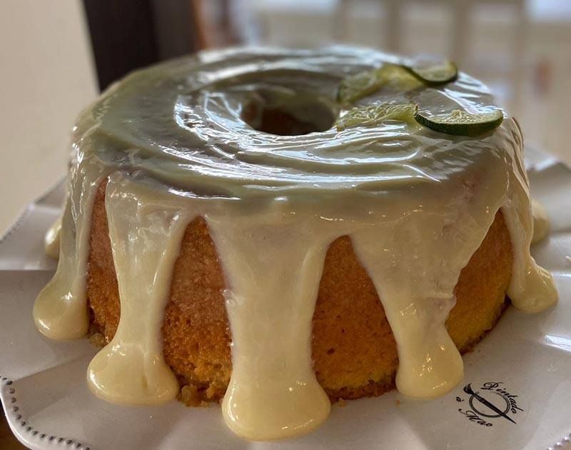 Um bolo redondo dourado, coberto com uma espessa camada de glacê claro e decorado com finas fatias de limão, servido num prato branco.