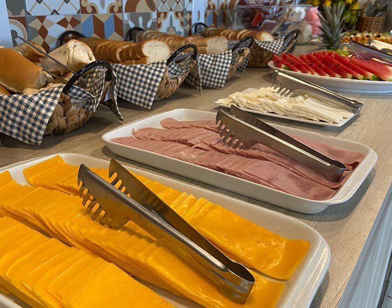 Um buffet de café da manhã com travessas de queijo fatiado, presunto, pão branco e melancia em um balcão de azulejos estampados.