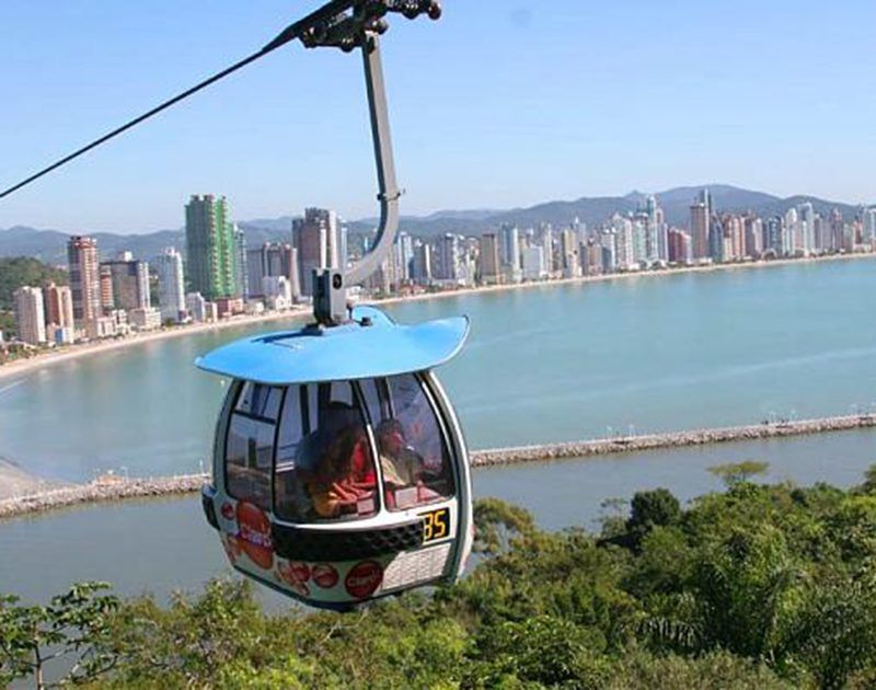 Um teleférico azul, número 35, suspenso sobre colinas verdejantes com o horizonte da cidade e a praia ao fundo.