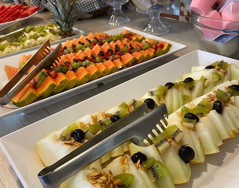 Um bufê com bandejas de melão fatiado, kiwi e uvas, com pegadores de metal para servir nas travessas.