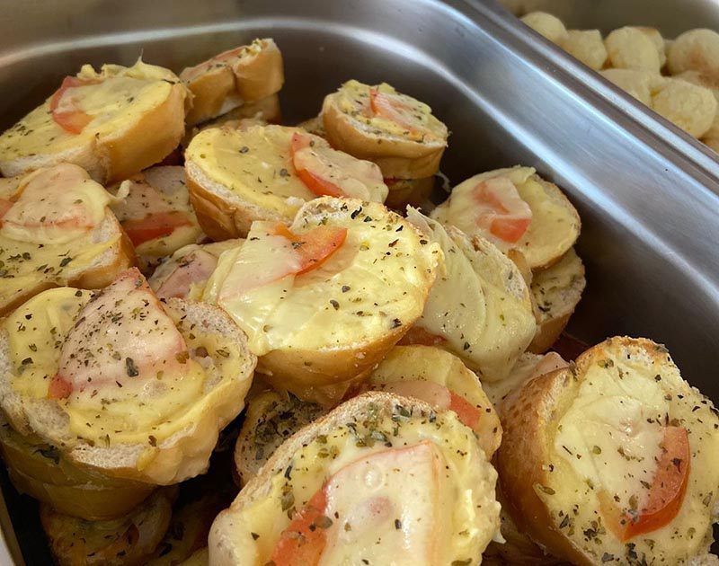 Uma bandeja de metal repleta de fatias de baguete tostadas, cobertas com queijo derretido, rodelas de tomate e ervas secas.