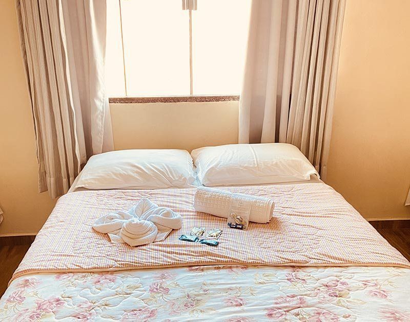 Uma cama de casal com travesseiros brancos e uma colcha floral rosa, além de uma toalha em formato de cisne, lençóis dobrados e pequenas comodidades.