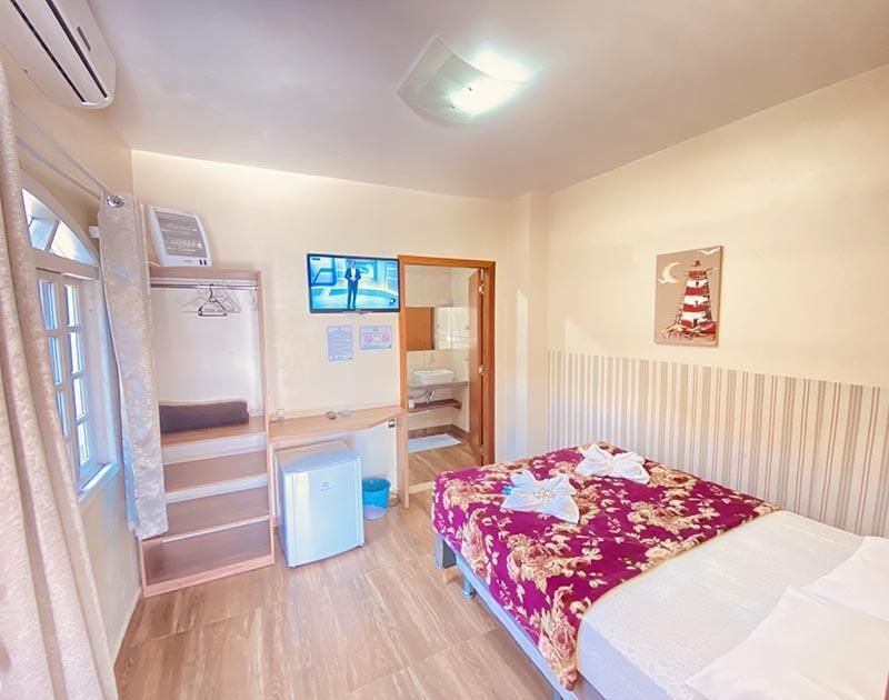 Um quarto de hotel limpo com uma cama com colcha rosa floral, uma escrivaninha com TV e frigobar, e uma porta de banheiro aberta.
