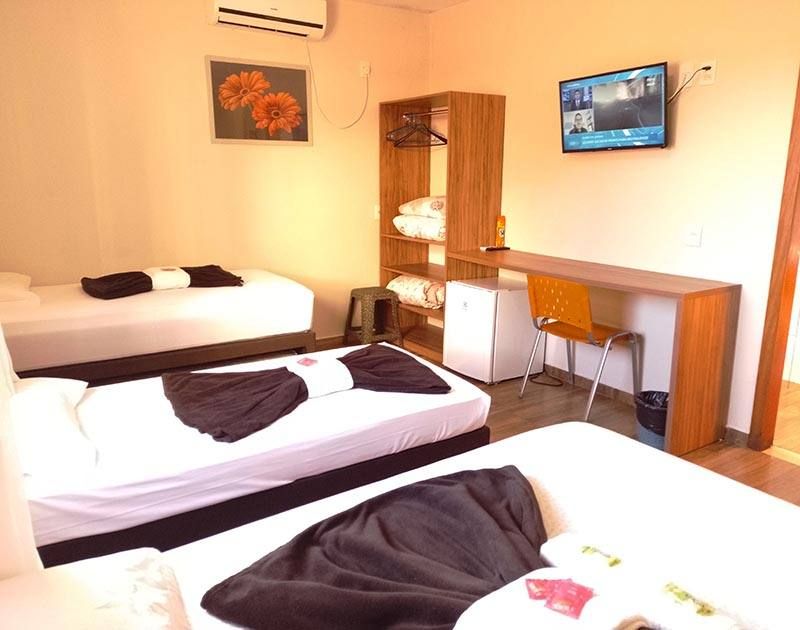 Um quarto de hotel com três camas com lençóis brancos e cobertores escuros, uma prateleira de madeira, uma escrivaninha, uma cadeira e uma TV.