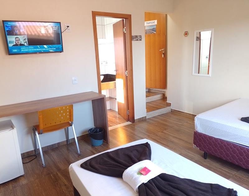 Um quarto de hotel com duas camas com toalhas marrom-escuras, uma TV fixada na parede, uma escrivaninha com cadeira e a porta do banheiro aberta.