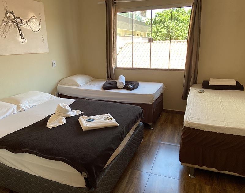 Um quarto de hotel com uma cama de casal à esquerda, uma cama de solteiro no centro ao fundo e uma cama de solteiro à direita.