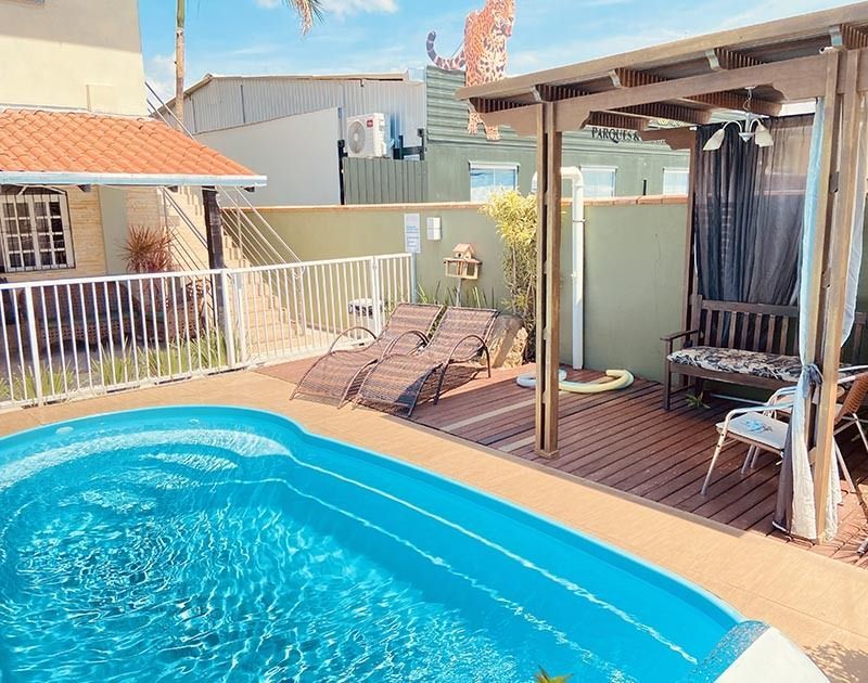 Uma piscina azul vibrante fica ao lado de um deck de madeira com espreguiçadeiras e uma área de pátio coberta por uma pérgola.