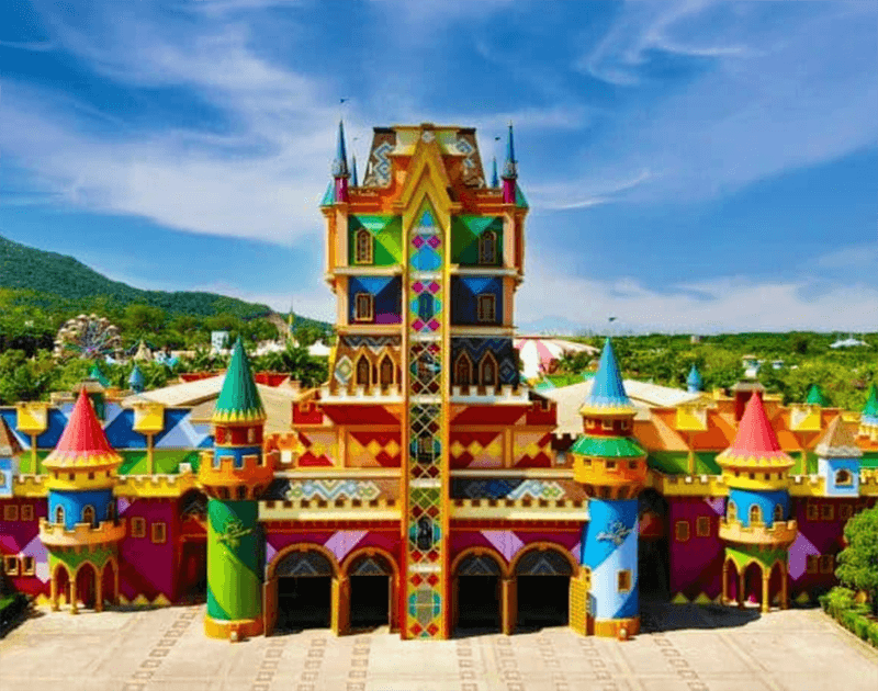 Um castelo colorido e simétrico com padrões geométricos vibrantes, torres e entradas em arco, situado num parque ensolarado ao ar livre.