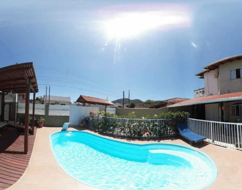 Uma piscina ao ar livre rodeada por um pátio, uma pérgola de madeira e uma casa sob um céu claro e ensolarado.