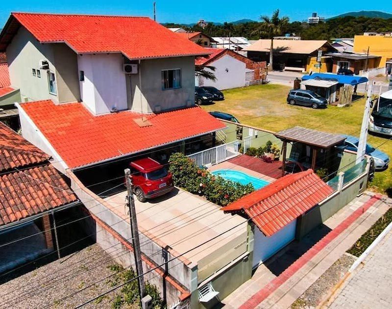 Uma casa de dois andares com telhado vermelho, piscina e um carro vermelho estacionado na entrada da garagem em um dia ensolarado.