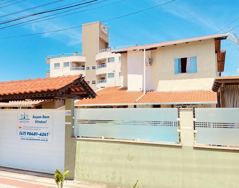 Um prédio de vários andares e uma casa vistos da rua, atrás de uma cerca branca com uma placa de imobiliária.