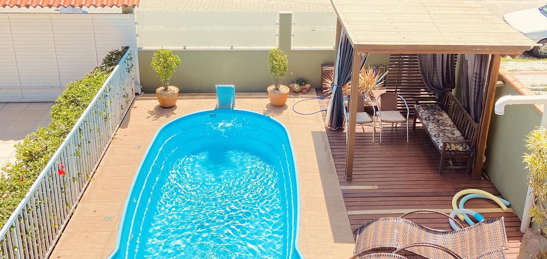 Uma piscina oval de um azul vibrante está localizada em um pátio ao lado de uma pérgola de madeira com cadeiras e um banco.