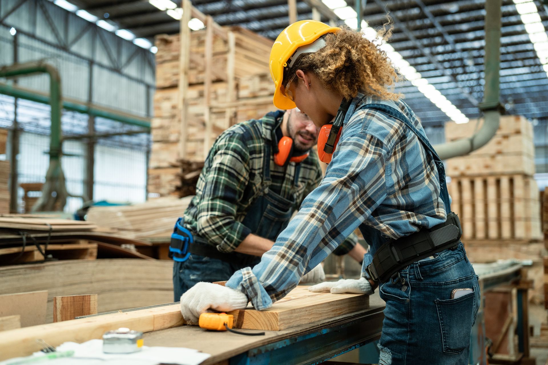 Two workers in hard hats measuring wood at a workbench in a lumber warehouse