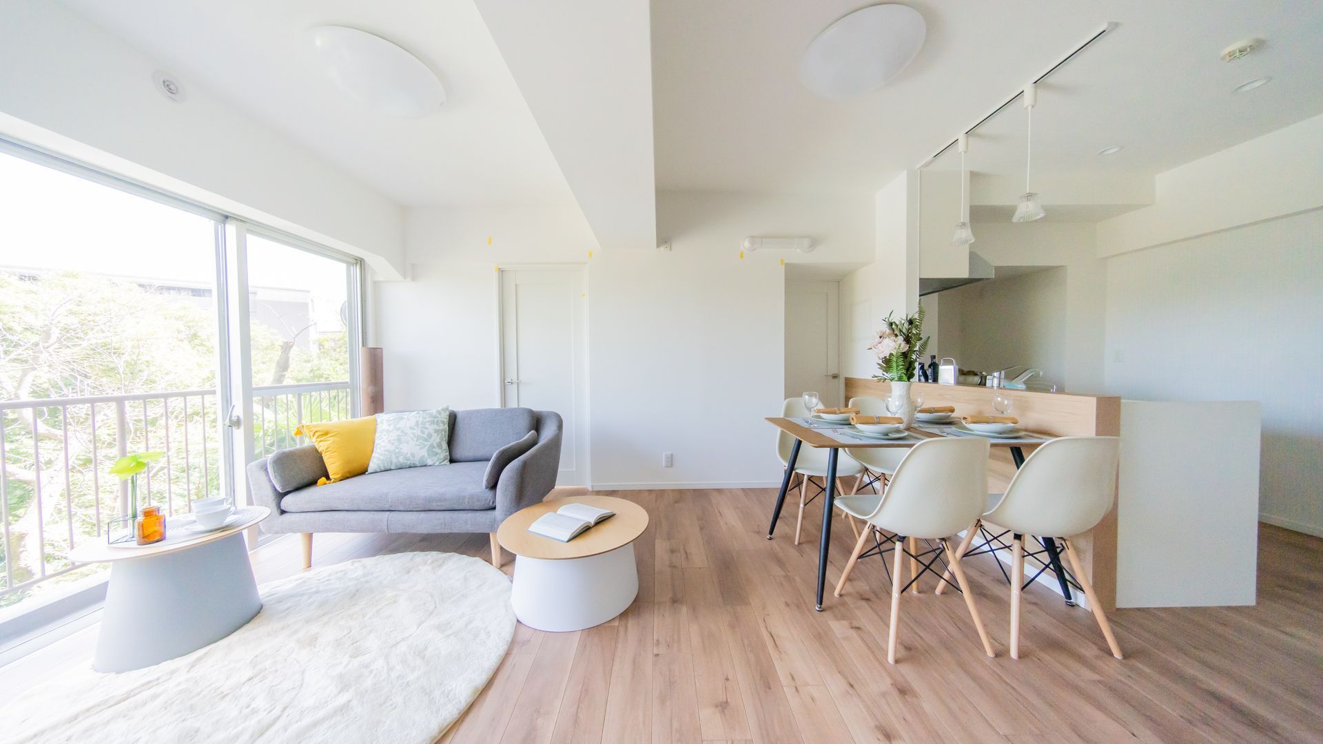 Bright modern living-dining room with gray sofa, round coffee table, and white dining set by large windows.