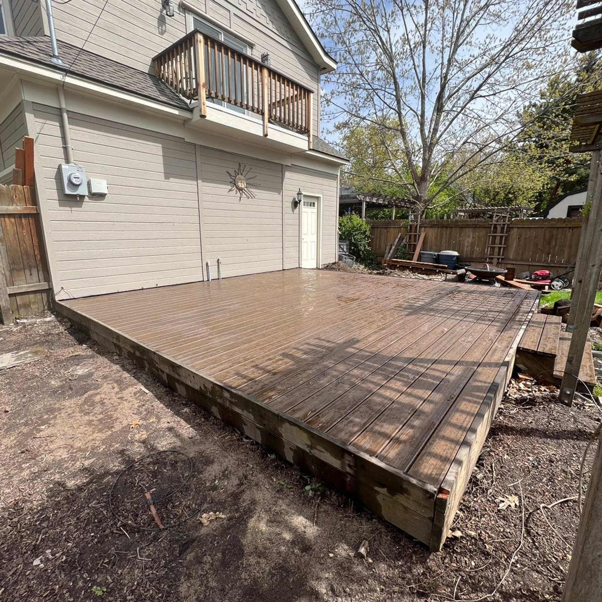Backyard deck beside a beige house, with railing, stairs, and a fenced yard under spring trees