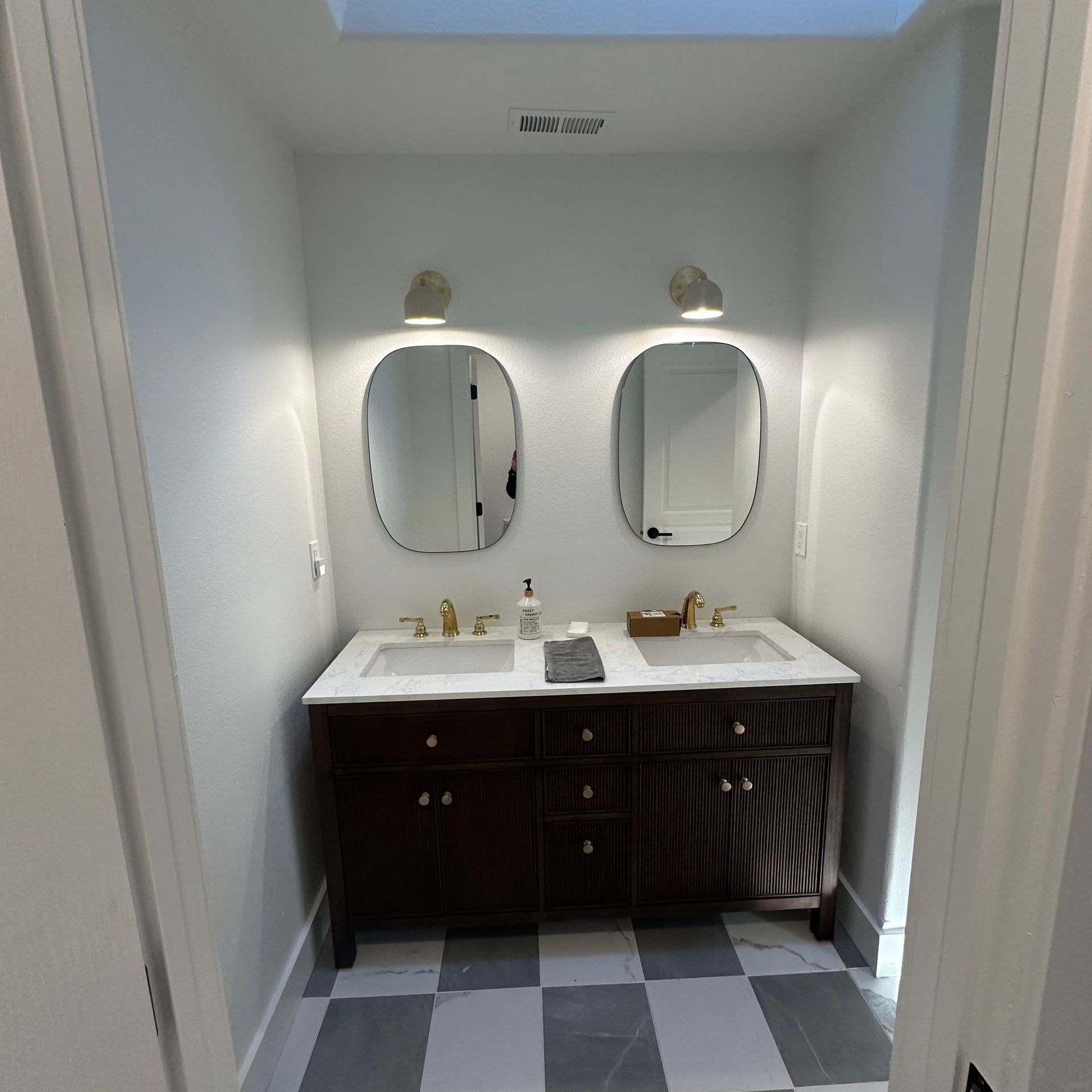 Small bathroom vanity with two mirrors, white counters, dark cabinets, and checkered floor.
