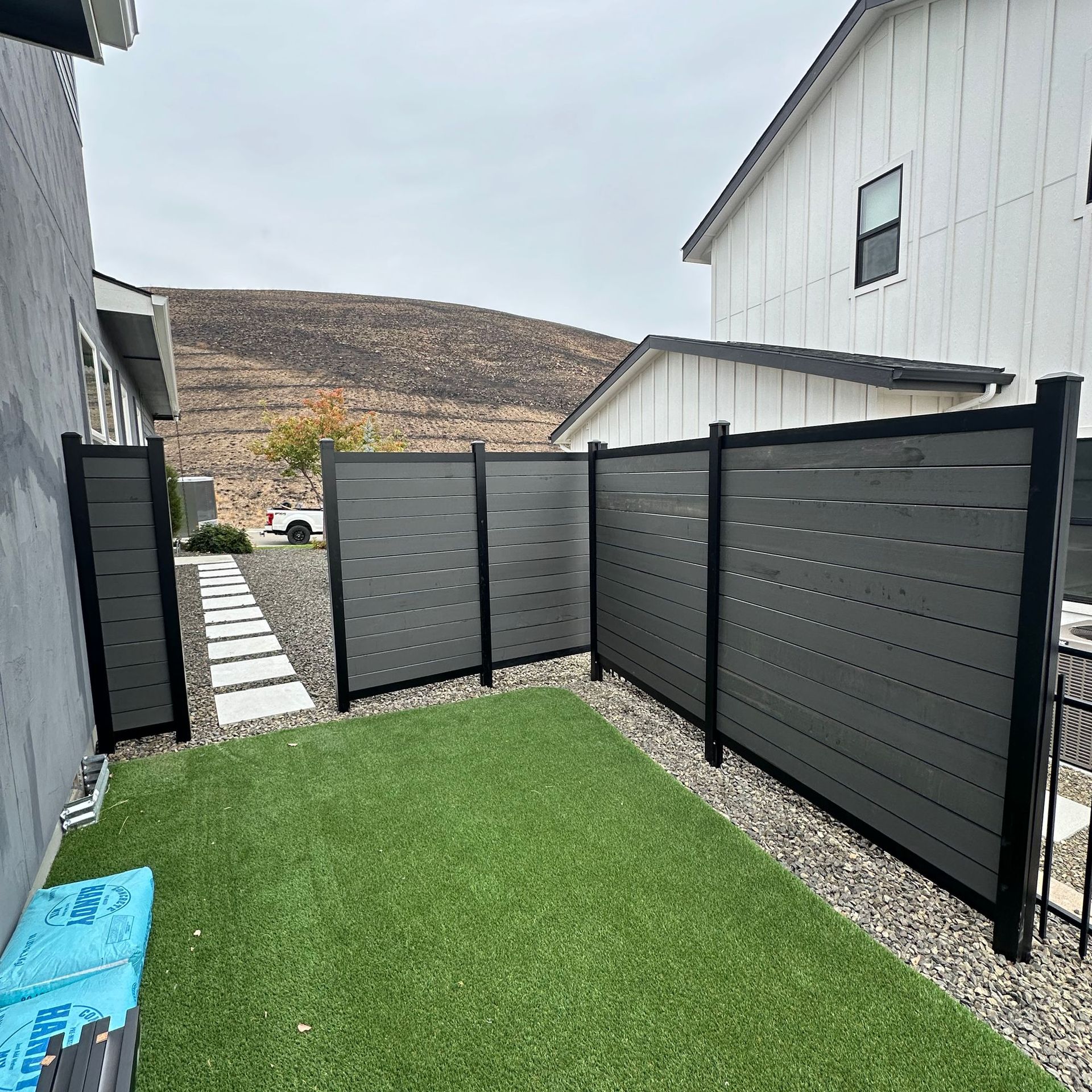 Narrow fenced backyard with artificial grass, gravel border, and gray privacy panels beside a house.
