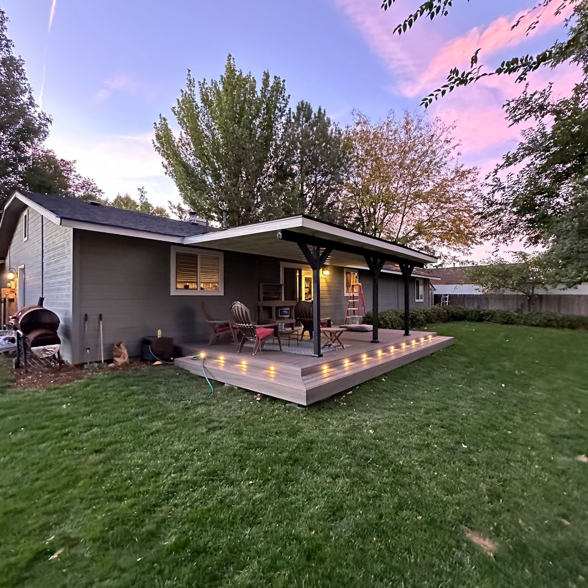 Backyard patio with a covered deck, string lights, chairs, and a grassy lawn at sunset.