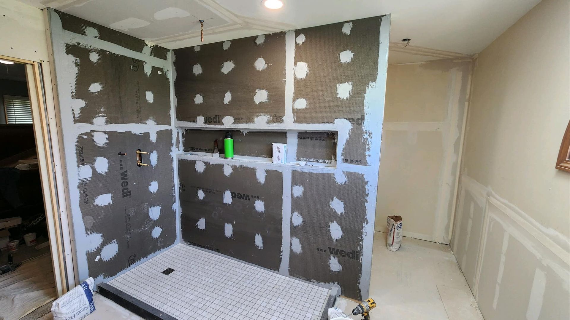 Bathroom shower area under renovation, with tiled walls, white trim, and a green bottle on a shelf.