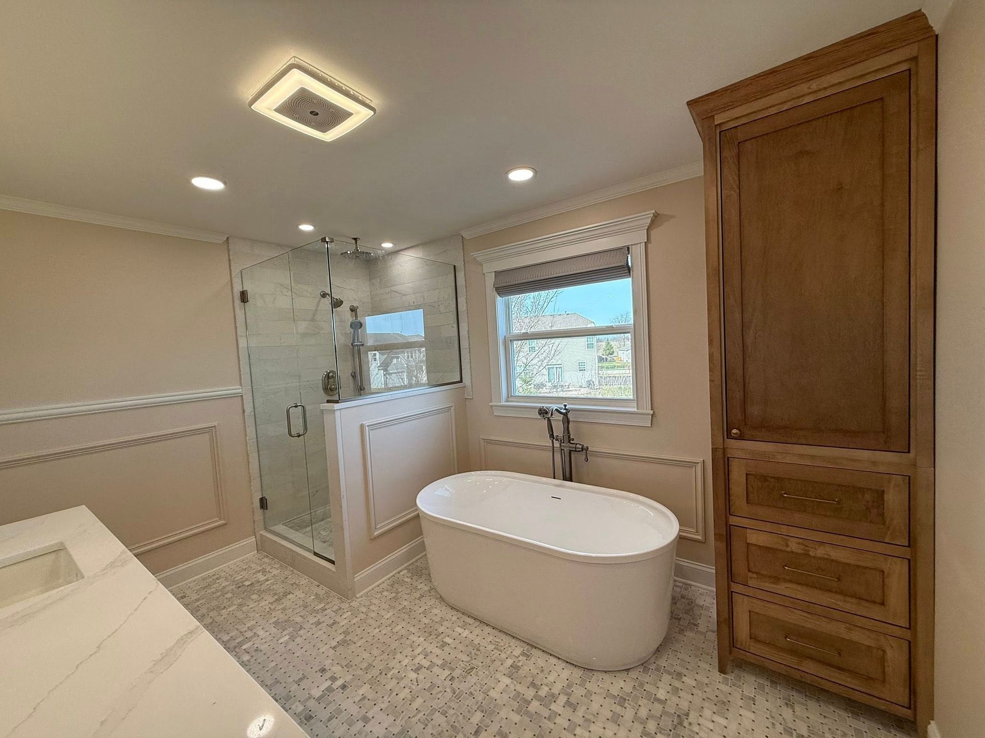 Bright modern bathroom with a freestanding tub, glass shower, window, and tall wooden cabinet.