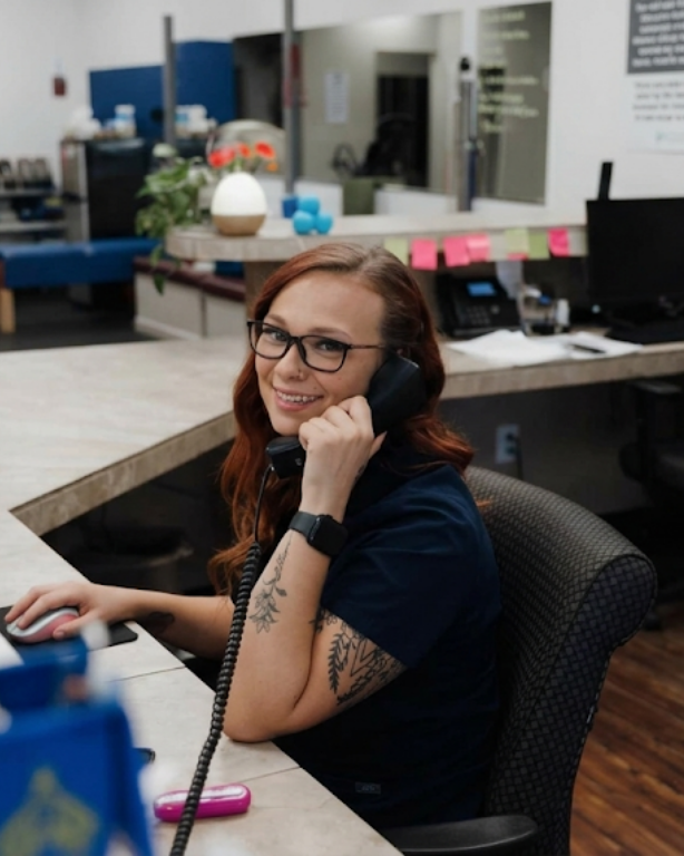 Woman at a desk in an office, smiling while talking on a corded phone with a computer nearby.